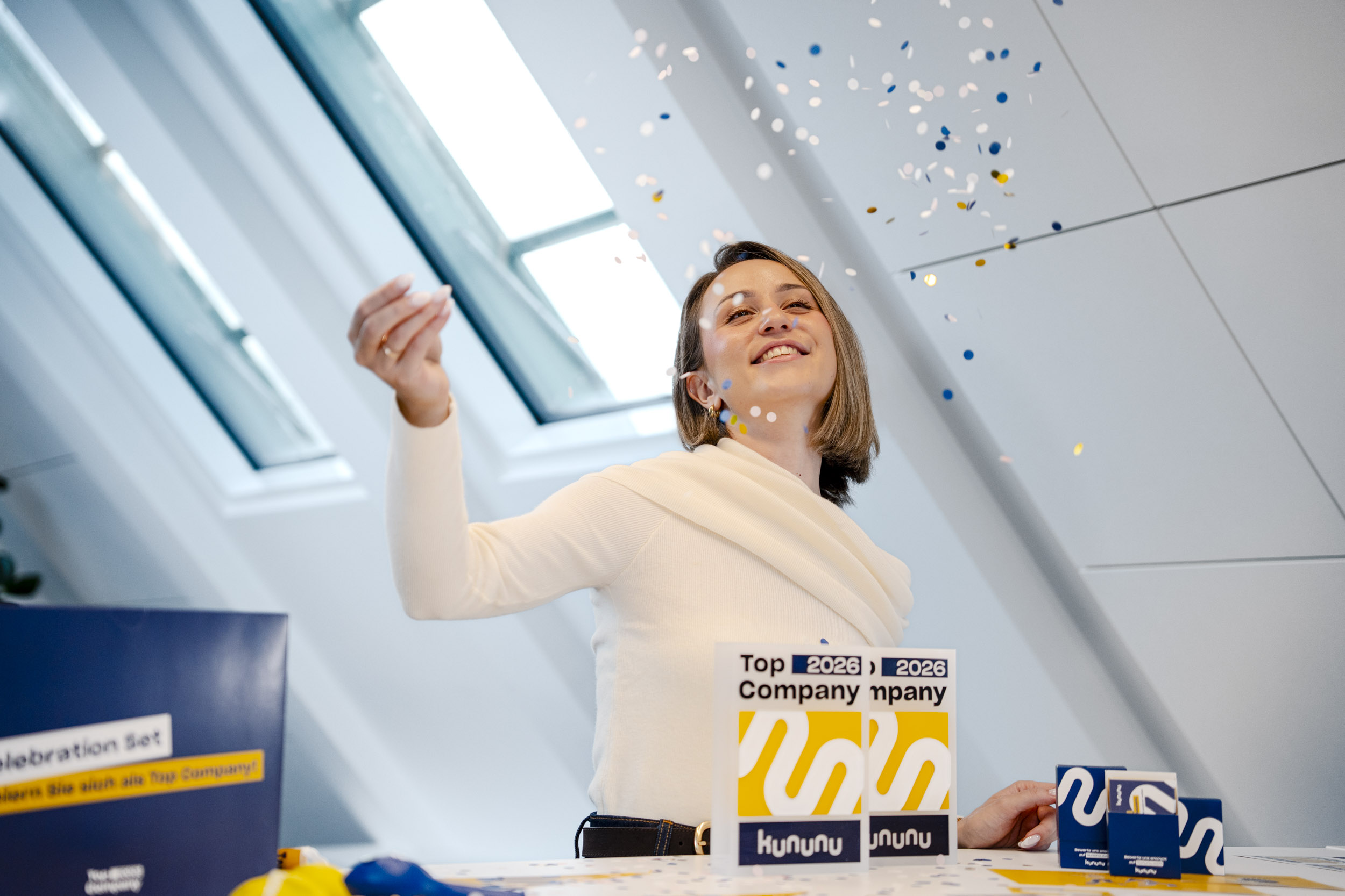 Abgebildet ist eine freudige Kollegin, die die kununu Top Company 2026 Box ausgepackt hat und das Konfetti des neuen Party-Sets in die Luft wirft