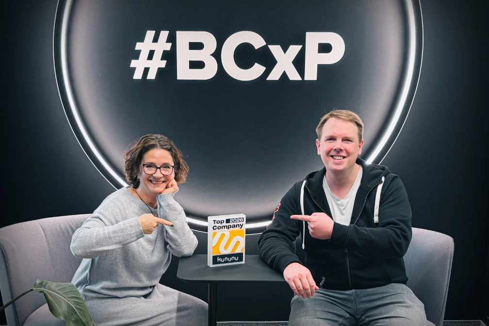 Zwei Personen sitzen vor Wand mit #BCxP und zeigen auf Kununu Auszeichnung Top Company 2026.