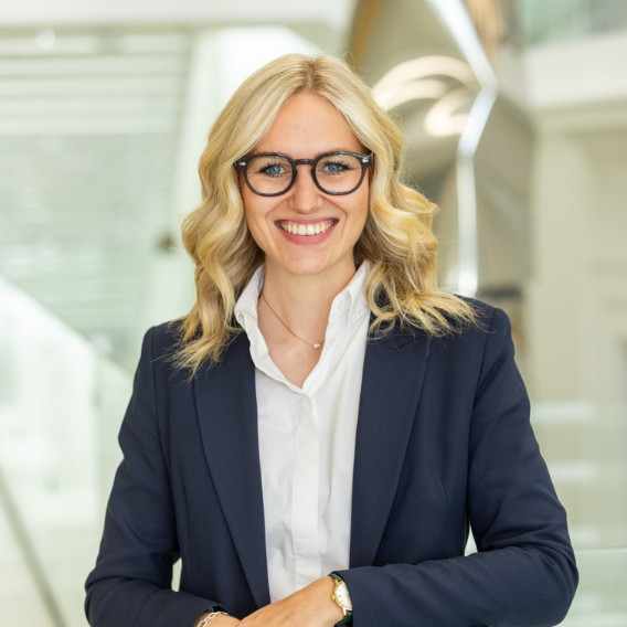 Blonde Frau mit Brille, weißem Hemd und dunkelblauem Sakko vor moderner Architektur.