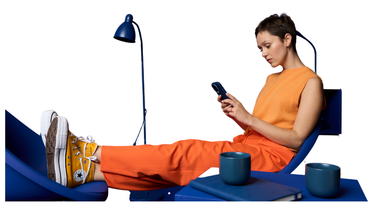 Person entspannt auf blauem Stuhl, orange Outfit, gelbe Sneakers, mit Smartphone, Tisch mit Büchern.