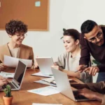 Menschen, die um einen Tisch herum vor ihren Laptops sitzen und motiviert zusammen arbeiten.