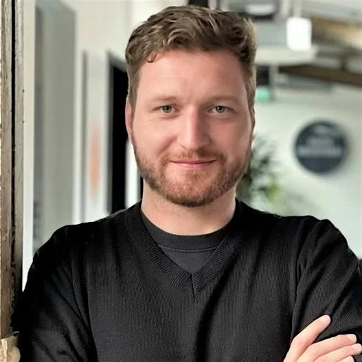 Christian Schlodder von der Suchmeisterei GmbH ist ein Mann mit kurzem Haar und Bart in schwarzem Pullover. Er steht mit verschränkten Armen vor einem Büroflur mit Pflanzen und Glaswänden.