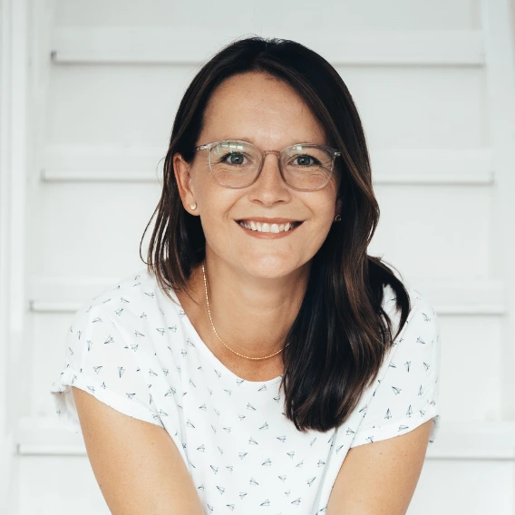 Lächelnde Frau mit Brille, dunklem Haar und weißen T-Shirt vor hellem Hintergrund.
