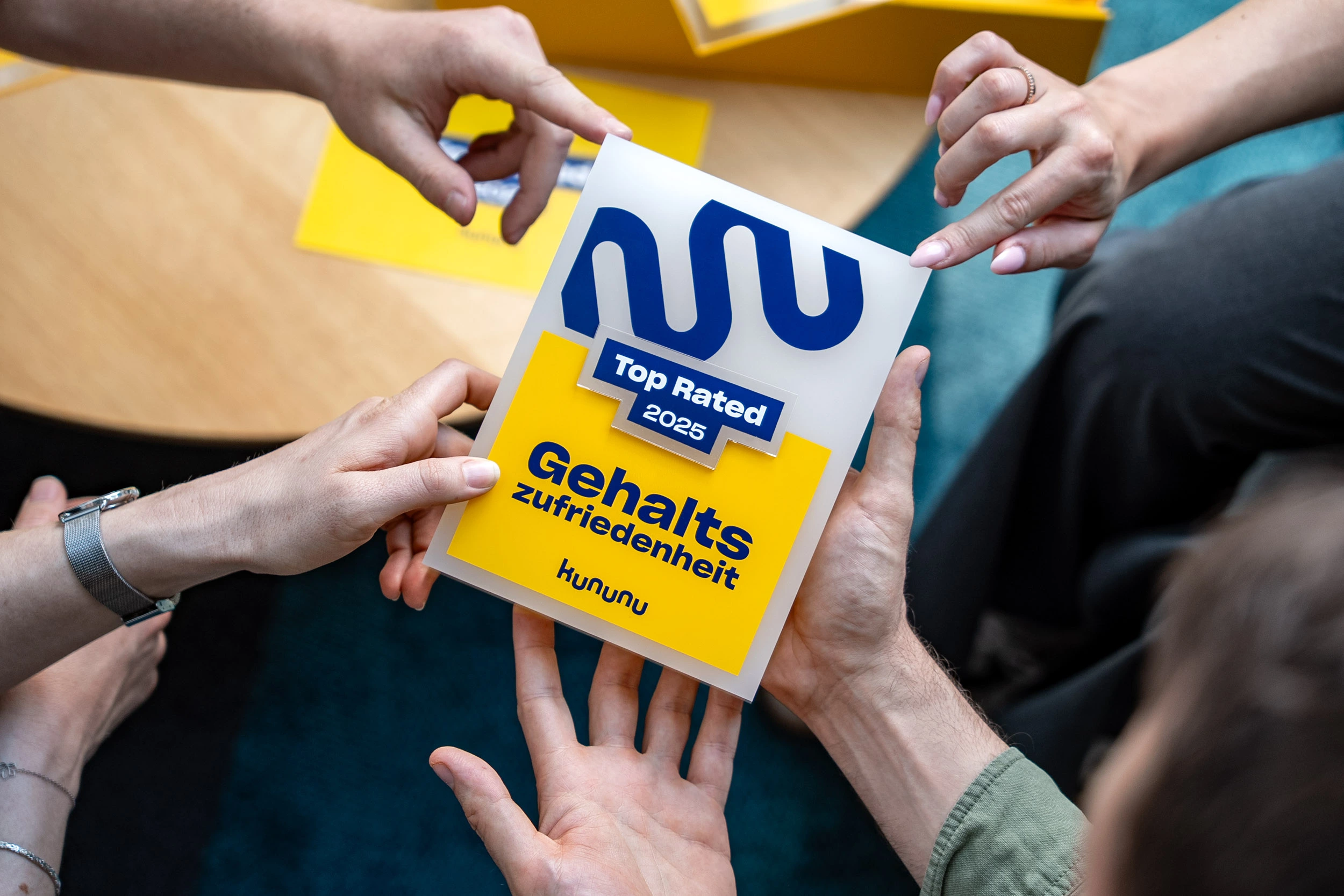 Das gelbe kununu Arbeitgebersiegel "Top Rated 2025 -Gehaltszufriedenheit", das von mehreren Händen gehalten wird.